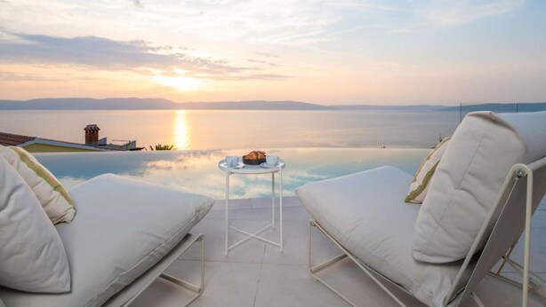A terrace with two white cushioned chairs, a small round table with a basket and cups, and a view of the sunset over a calm sea. - Amazing Villa in Labin With Sauna, Private Pool (Rasa)