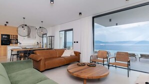 A modern living room with a large window, a sofa, a coffee table, and a dining area. - Amazing Villa in Labin With Sauna, Private Pool (Rasa)