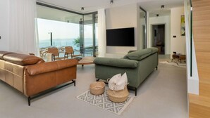 A modern living room with a leather sofa, a green sofa, and a dining area with chairs. There is a TV mounted on the wall and a large window offering a view of the outdoors. - Amazing Villa in Labin With Sauna, Private Pool (Rasa)