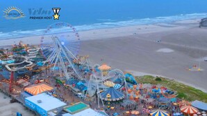 Luftaufnahme eines Freizeitparks direkt am Strand mit einem großen Riesenrad, verschiedenen Fahrgeschäften und Sitzgelegenheiten.