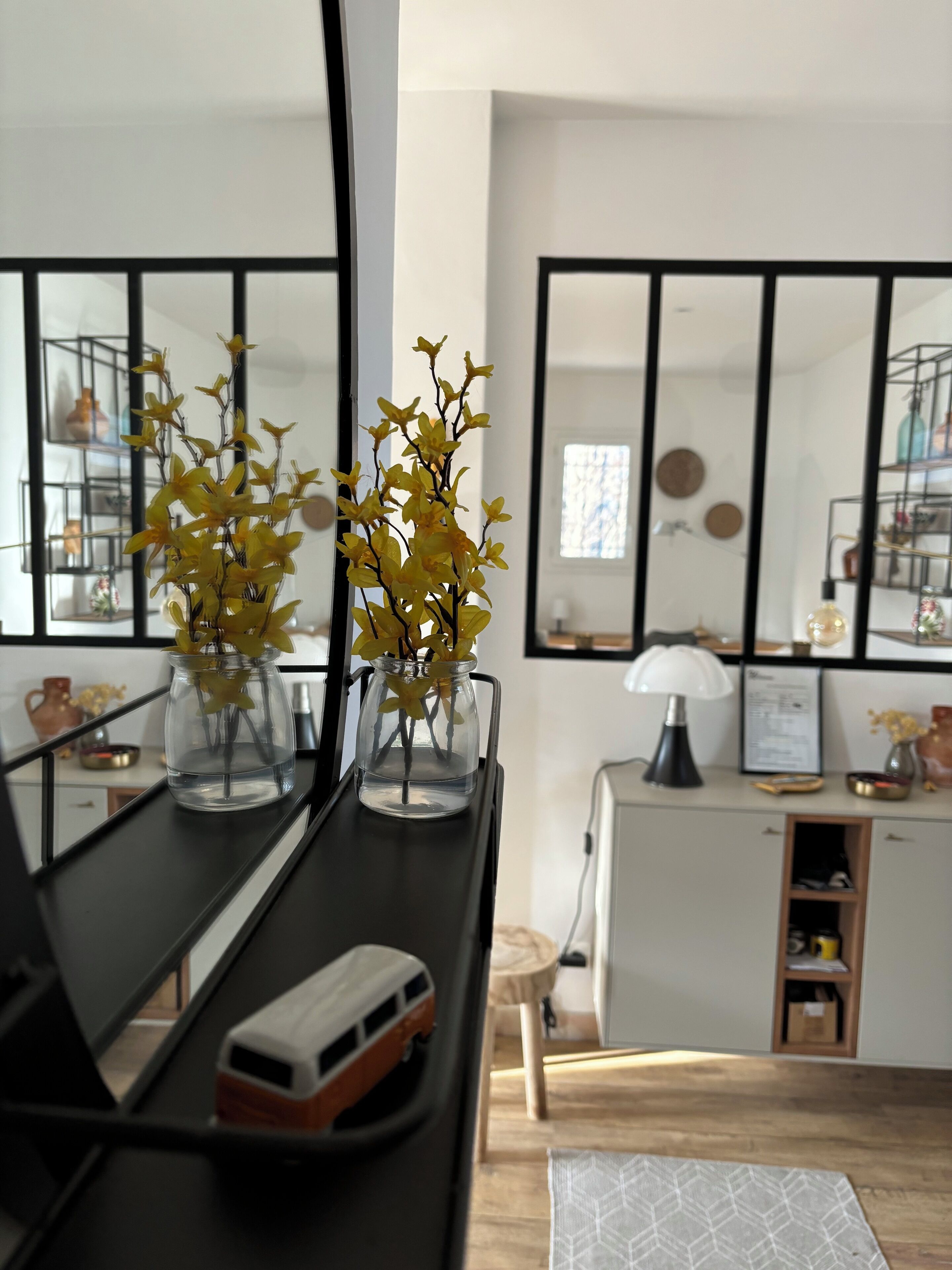 A kitchen with a black countertop, two glass vases with yellow flowers, a white cabinet, and a mirror above the counter.