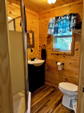 Une salle de bain rustique, aménagée dans une cabane en bois, avec un lavabo, des toilettes et une fenêtre donnant sur la forêt.