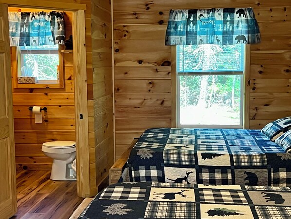 L’intérieur d’une cabane en bois comprend un lit, une fenêtre donnant sur la forêt, des toilettes et un rideau orné de motifs animaliers.