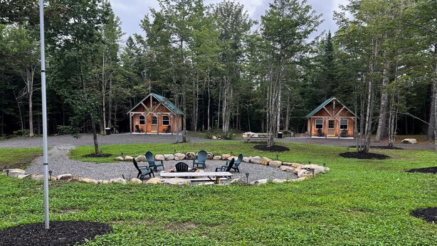 Two wooden cabins with green roofs, surrounded by a gravel area with green chairs and a fire pit, set in a forested area. - Cabin 2 Recreational Cozy Cabin Rentals (Sangerville)
