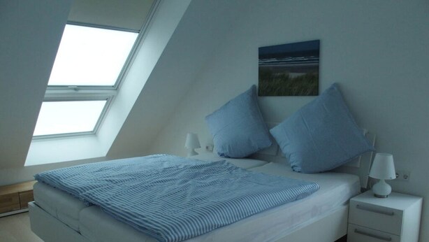 A bedroom with a skylight, a bed with blue bedding, a nightstand with a lamp, and a wall-mounted picture. - Lutje Sandbank Modern Retreat (Juist)