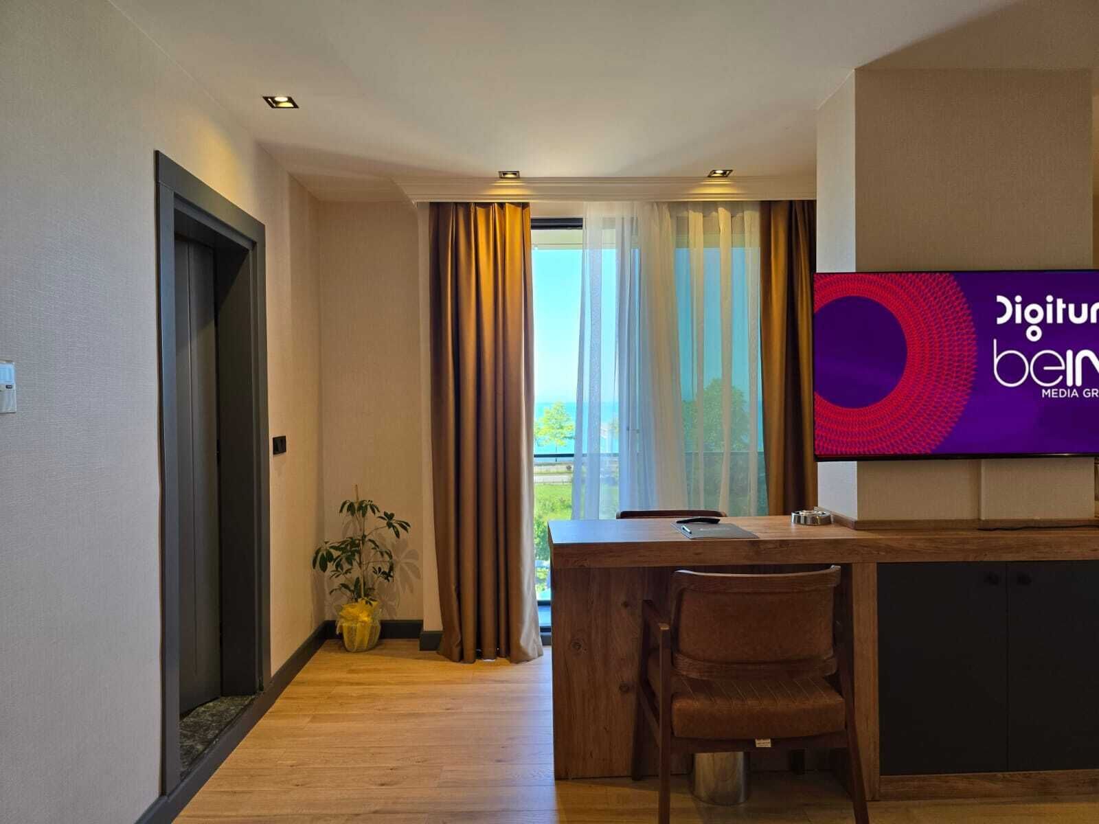 A modern hotel room with a wooden floor, a desk with a chair, a large TV screen, and a view of the outdoors through a window with curtains.