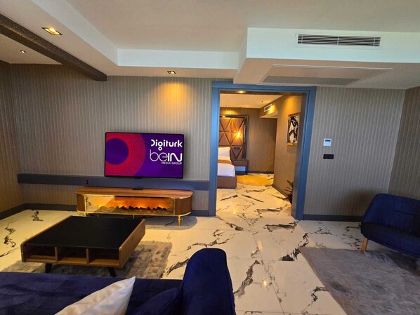 Un moderno vestíbulo de hotel con un televisor de pantalla plana mostrando 'Digiturk bein MEDIA GROUP', una chimenea, un sofá y una alfombra estampada.
