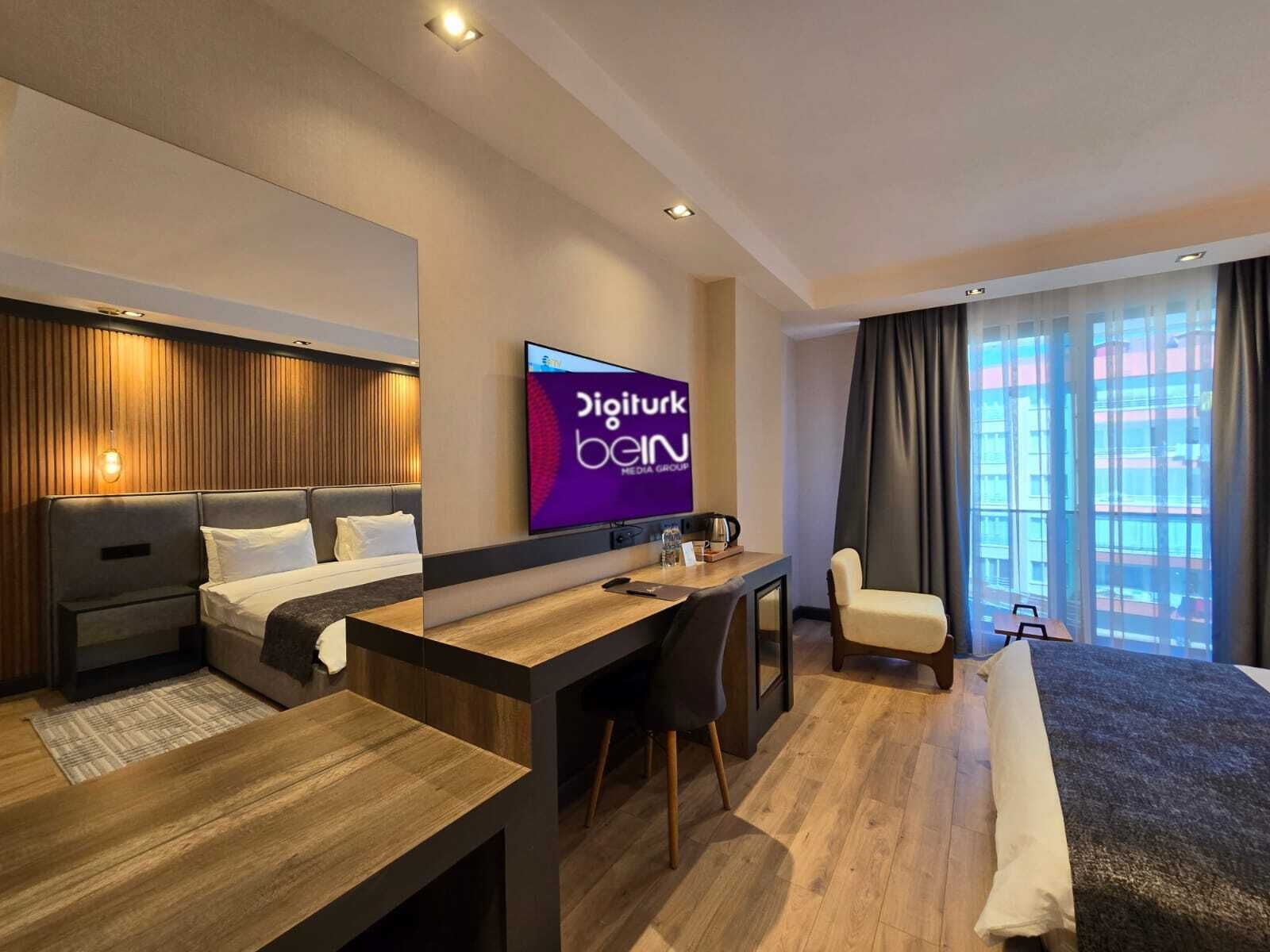 A modern hotel room with a bed, a desk, a chair, and a television displaying 'Digiturk Bein'.