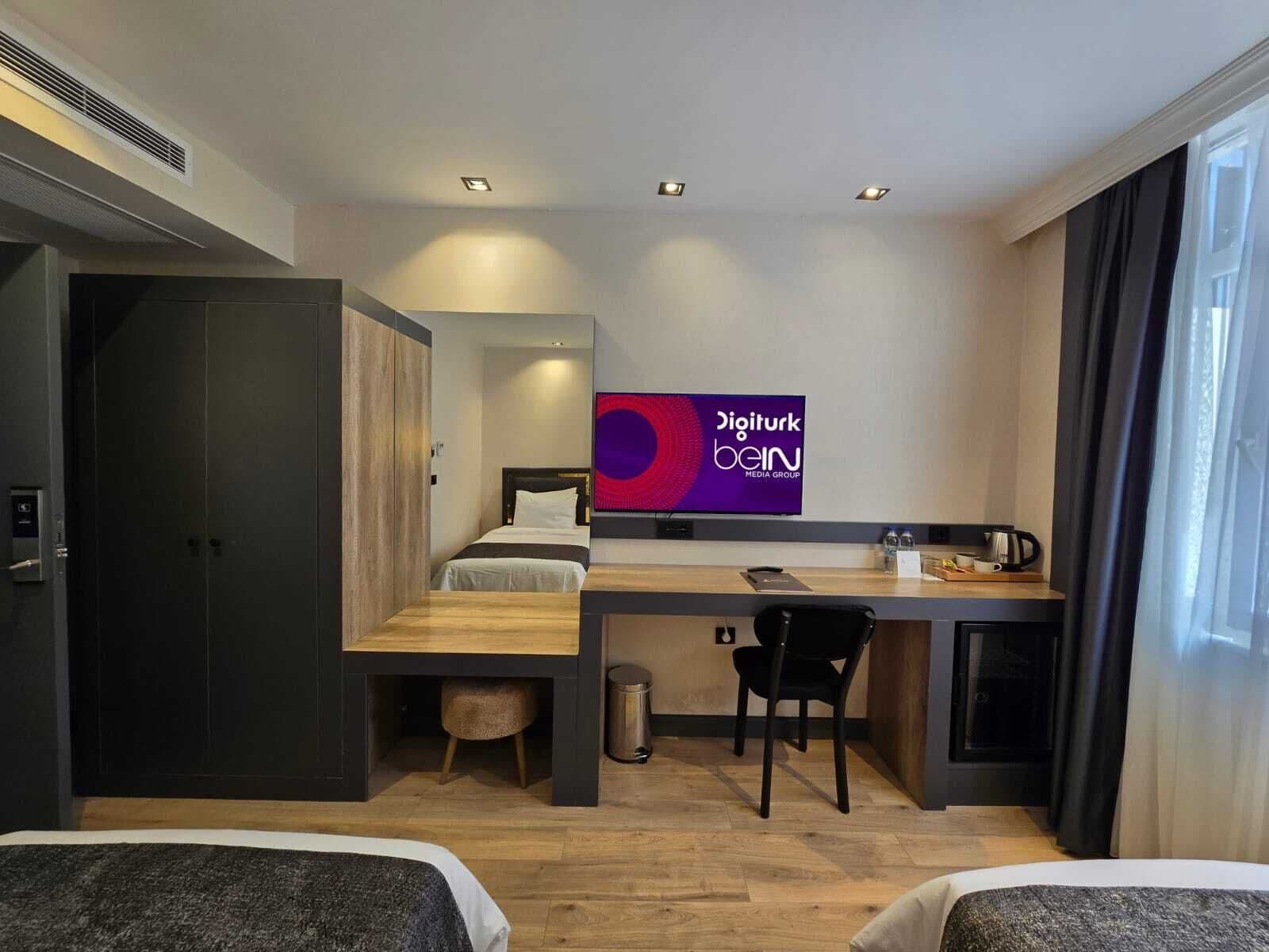A modern hotel room with a bed, desk, and chair, a TV displaying 'Digiturk' branding, and a window with curtains.