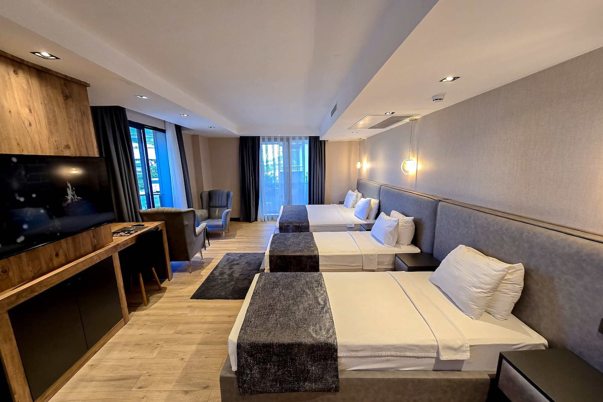 Ein modernes Hotelzimmer mit Flachbildfernseher, Schreibtisch, zwei Betten, einer Couch und einem großen Fenster mit Vorhängen.