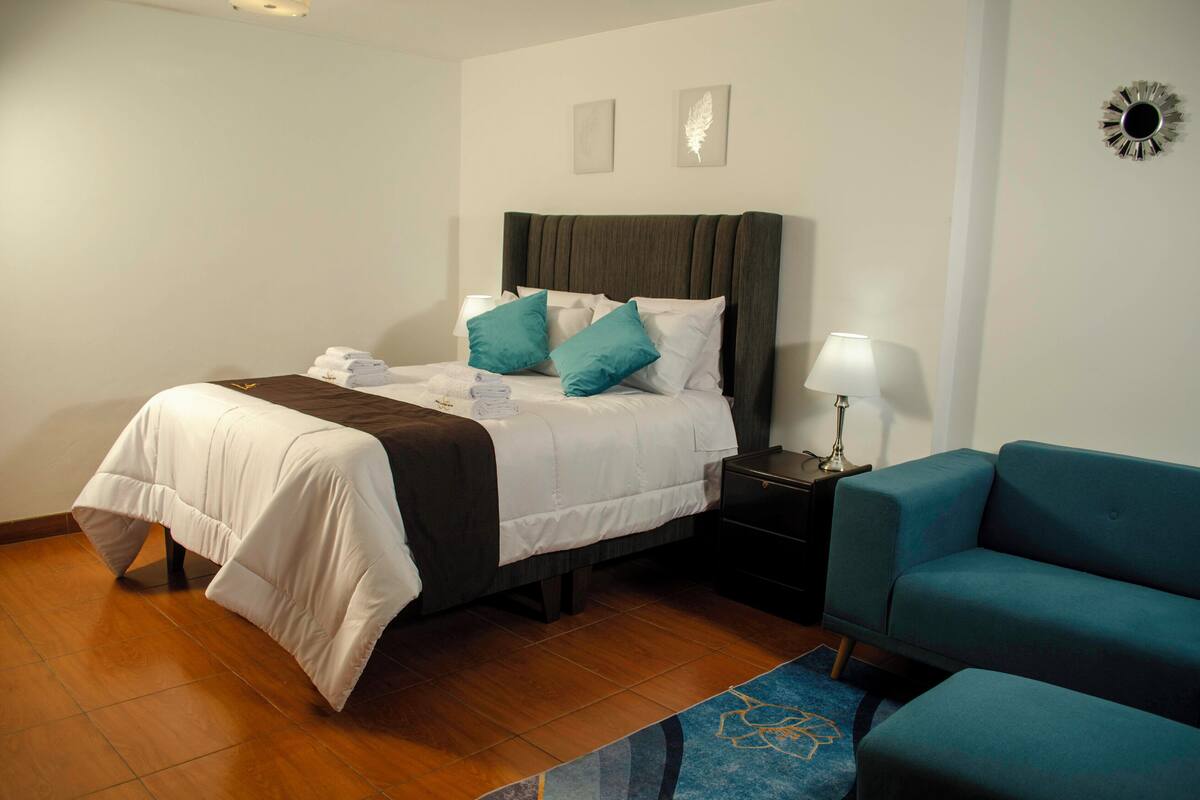 A neatly made bed with a white comforter and teal pillows, a blue sofa, and a small bedside table with a lamp.