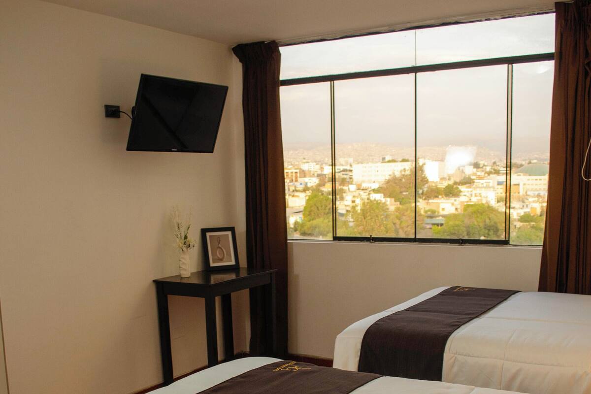 A hotel room with two beds, a flat-screen TV mounted on the wall, a small table with a vase and picture frame, and a window with a city view.
