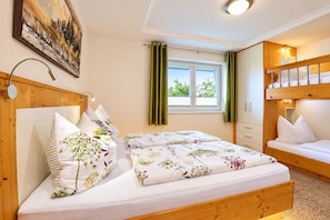 Une chambre avec une tête de lit en bois, un lit recouvert d’une literie à motifs floraux, une fenêtre avec des rideaux et un lit superposé avec des oreillers.