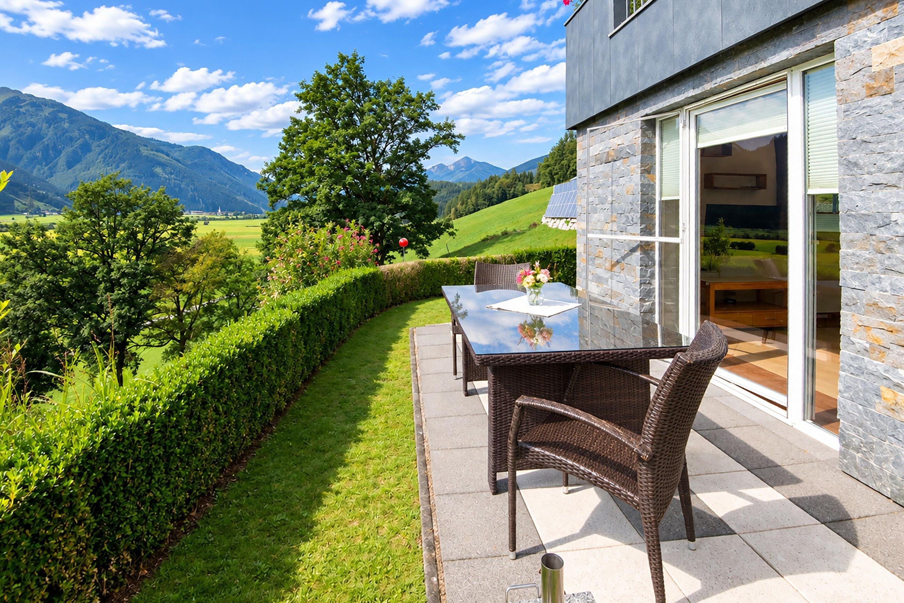 Une terrasse avec une table et des chaises, une porte vitrée et une vue sur un paysage verdoyant avec des montagnes.