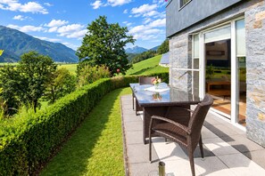 Une terrasse avec une table et des chaises, une porte vitrée et une vue sur un paysage verdoyant avec des montagnes.