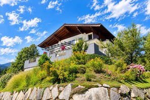 Une maison à deux étages avec un toit en pente, un balcon doté d’une balustrade en métal et un mur de pierre en premier plan.