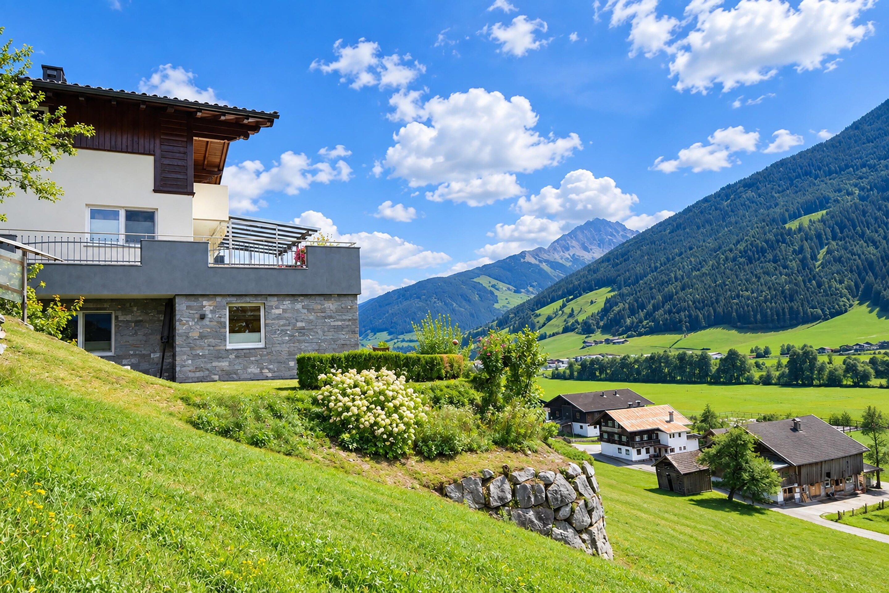 A modern two-story house with a balcony, surrounded by lush greenery and mountains.