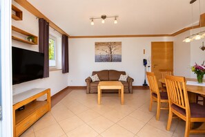 A living room with a flat-screen TV, a brown sofa, a wooden coffee table, and a dining area with wooden chairs and a table. - Apartment in Mittersill Near ski Lift (Mittersill)