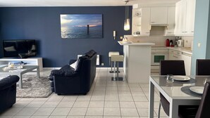A modern living room with a dark blue wall, a flat-screen TV, a black leather sofa, and a glass coffee table.