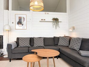 A modern living room with a sectional sofa, two wooden side tables, a hanging wicker light fixture, a framed abstract artwork, a wall-mounted shelf with decorative items, and a floor lamp. - 4 Star Holiday Home in Aabenraa (Aabenraa)