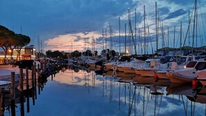 A marina with boats docked at wooden piers, calm water reflecting the boats and sky, and buildings with lights on in the background. - Holiday Home Near Spiaggia Tartaruga (Caorle)