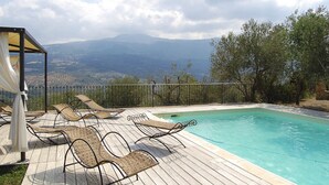 Une piscine entourée de chaises longues et agrémentée d’une pergola, le tout surplombant une terrasse en bois et offrant une vue sur les montagnes lointaines.