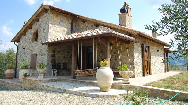 Une maison en pierre avec un toit en tuiles, une cheminée et un porche doté d’une balançoire.