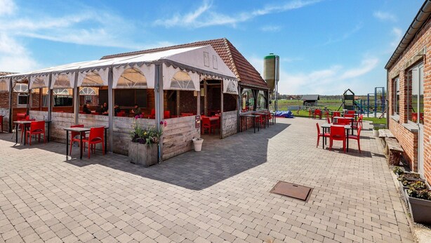 An outdoor café with brick tables and red chairs, a covered seating area with a white canopy, and a playground in the background. - Cottage on Sheep Farm With Gelateria (Merksplas)