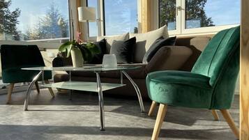 Ein modernes Wohnzimmer mit einem Glastisch, grünen Sesseln und einem Sofa mit vielen Kissen.