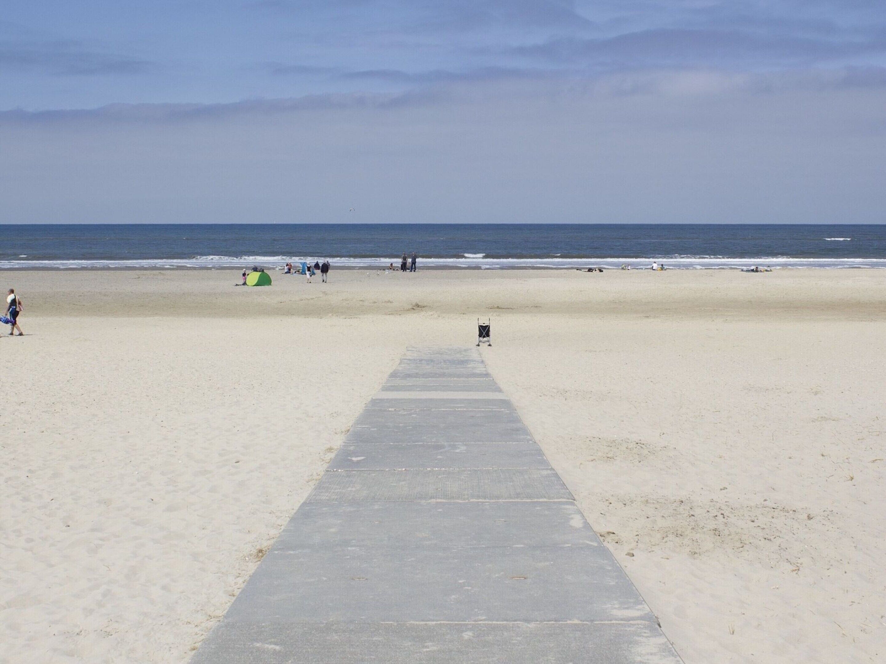 A beach with a concrete walkway leading to the water, a green tent, and people in the distance.