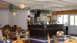 A restaurant with wooden tables and chairs, a bar area, and a view of the outdoors through large windows. - Holiday Room Lenzer Hafen (Fuenfseen)