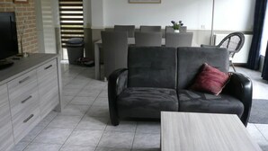 A modern living room with a grey sofa, a small table, and a TV on a cabinet. There is a dining area with chairs and a table, and a large window with blinds. - Holiday Apartment Zonnewende 16 (Sint Maartensbrug)