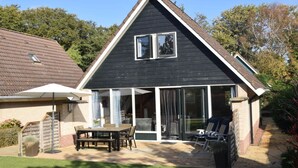 A two-story house with a dark exterior, a covered patio with a table and chairs, and a brick pathway leading to the house. - Holiday Apartment Zonnewende 16 (Sint Maartensbrug)