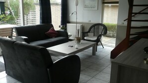 A modern living room with a sofa, a coffee table, and a chair. There is a staircase to the right and a window with curtains. - Holiday Apartment Zonnewende 16 (Sint Maartensbrug)