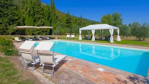 Piscina com espreguiçadeiras e toldo, rodeada por vegetação e uma colina.