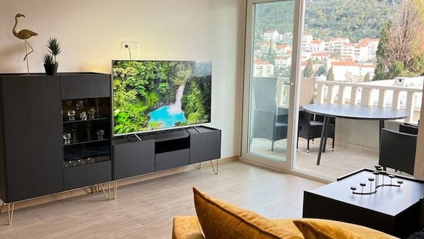 Un salon moderne doté d’un téléviseur à écran plat, d’un canapé, d’une table à manger et d’un balcon offrant une vue sur les bâtiments et les arbres.