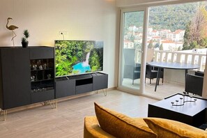 A modern living room with a flat-screen TV, a sofa, a dining table, and a balcony with a view of buildings and trees. - Apartments Jelena - One Bedroom Apartment With Balcony and Sea View (Dubrovnik)