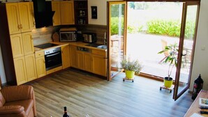 A compact kitchen with wooden cabinets, a microwave, and a stove. There is a dining area with a table and chairs, and a sliding glass door leading to an outdoor patio. - Rugen Apartment Waterview (Bergen auf Ruegen)