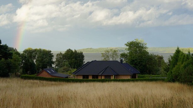 A house with a dark roof and brick walls, surrounded by tall grass and trees, with a lake and hills in the background. - Rugen Apartment Waterview (Bergen auf Ruegen)