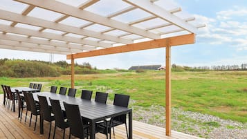 An outdoor covered patio with a long table and chairs, surrounded by a grassy landscape and a clear sky.