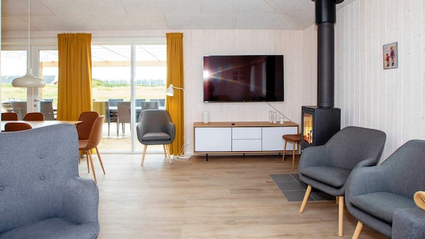 Un salon moderne doté d’un téléviseur à écran plat, d’un poêle à bois noir, de canapés gris et d’un coin repas agrémenté de rideaux jaunes.