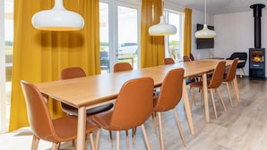 Un espace de salle à manger moderne avec une longue table en bois, des chaises orange, des rideaux jaunes et des luminaires suspendus.