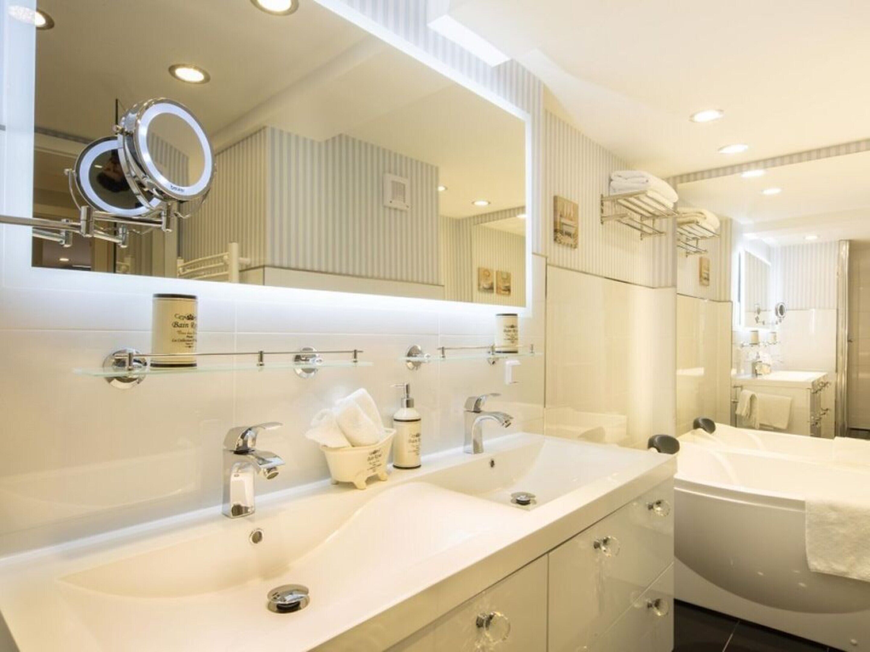 A modern bathroom with a large mirror, two sinks, a bathtub, and a glass shelf with toiletries.