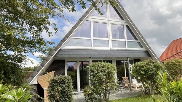 Et trekantet hus med en stor glasfacade, omgivet af grøn vegetation og en lille patio.