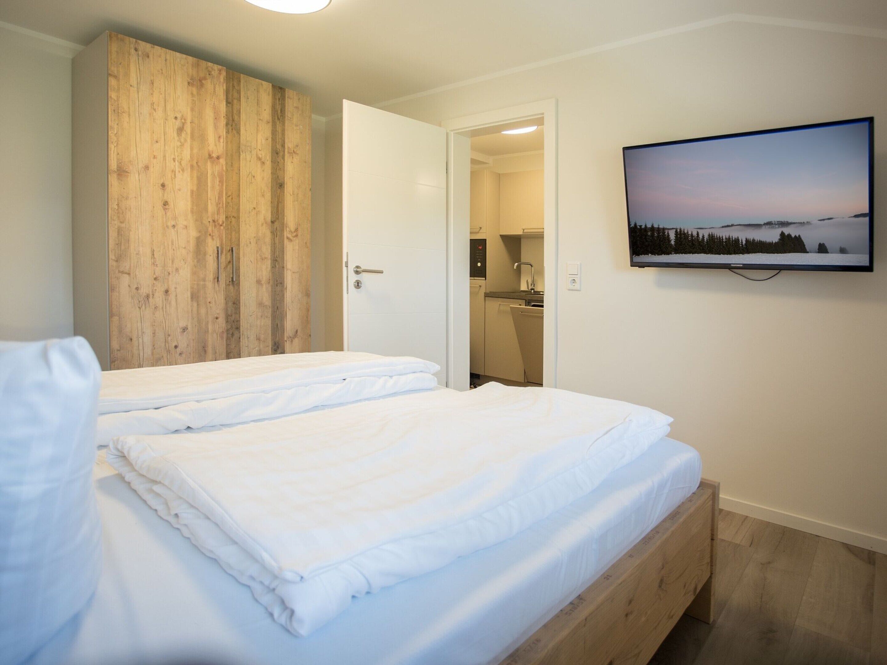 A bedroom with two beds, a wooden wardrobe, a TV on the wall, and a kitchen in the background.
