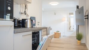 Une cuisine moderne équipée d’un four, d’un micro-ondes et d’un lave-vaisselle encastrés. On y trouve un meuble en bois et un petit tabouret en bois. Une plante est visible dans un coin.