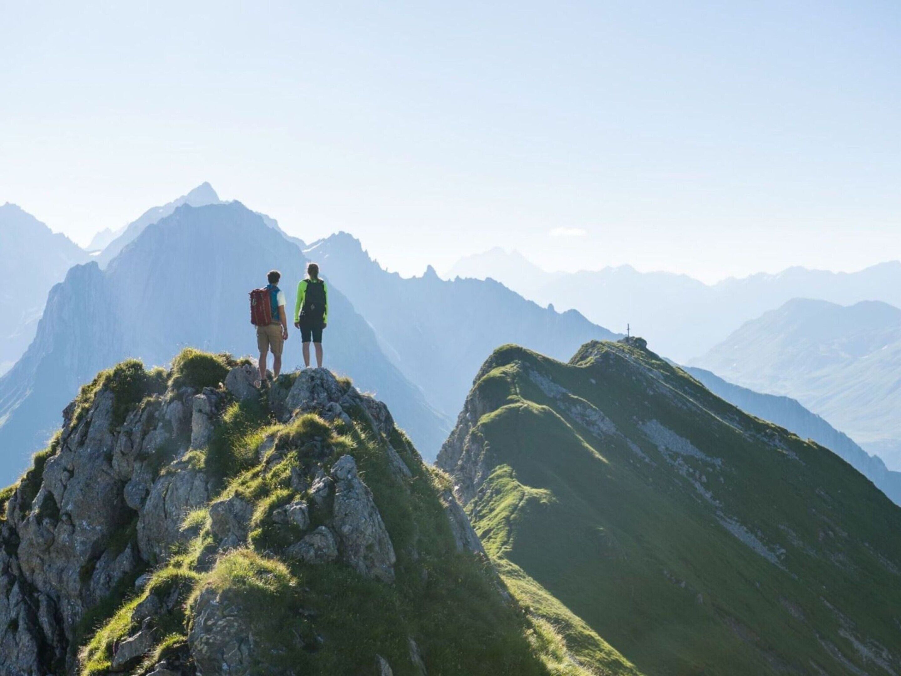 Deux personnes se tiennent au sommet d’une montagne, près d’une croix, entourées d’un paysage accidenté et de montagnes lointaines.