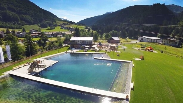 A large rectangular swimming pool with a diving platform, surrounded by a grassy area and buildings in a hilly landscape. - Flat in Arlberg Near Ski Slopes & Sauna (Klösterle am Arlberg)
