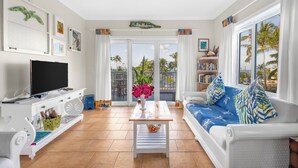 Ein Wohnzimmer mit Flachbildfernseher, weißen Möbeln, einem Couchtisch mit Blumen und Blick auf Palmen durch große Fenster.