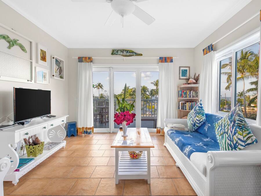Una sala de estar con una televisión de pantalla plana, muebles blancos, una mesa de centro con un jarrón de flores y vistas a palmeras a través de ventanas amplias.